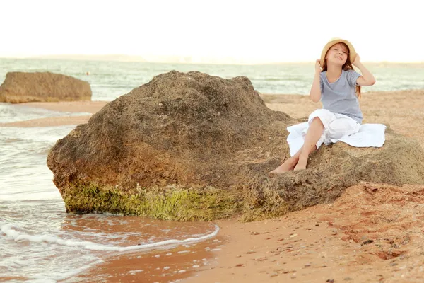 Cute little girl with long hair and healthy skin is sitting on a large rock near the sea. — Stock Photo, Image