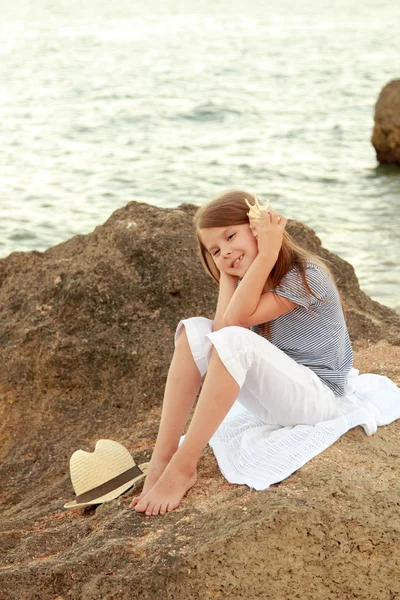 Glad söt liten flicka med ett vackert leende sitter på stranden på kvällen. — Stockfoto