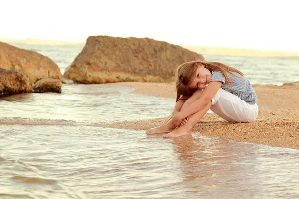 Positiva sonrisa niña en la playa moja los pies en el agua al atardecer en el aire libre . — Foto de Stock