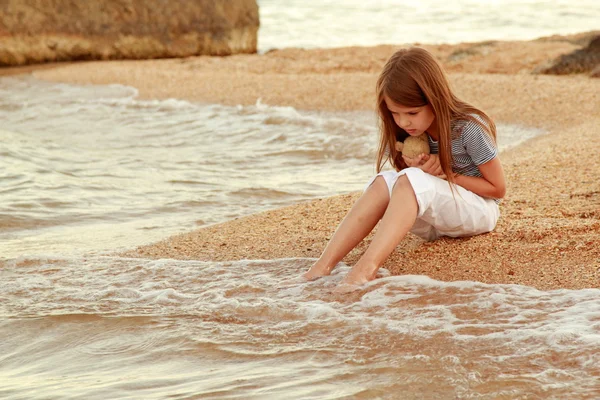 Gadis kecil tersenyum positif di pantai membasahi kaki dalam air saat matahari terbenam di luar ruangan . — Stok Foto