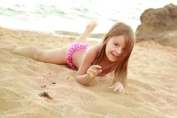 Schöne lächelnde kleine Mädchen in einem rosa Badeanzug wird im Sand am Strand gespielt — Stockfoto