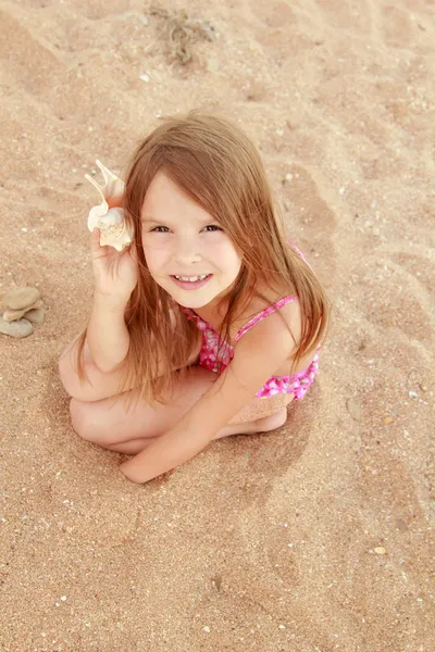 Europeo sorridente bambina in costume da bagno rosa seduta su uno sfondo di sabbia marina — Foto Stock