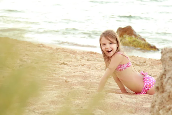 Красивая улыбающаяся маленькая девочка в розовом купальнике играет в песке на пляже — стоковое фото
