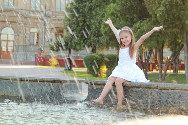 Veselé roztomilé děvčátko v bílých šatech sedí u fontány a usmívá se na letní téma — Stock fotografie