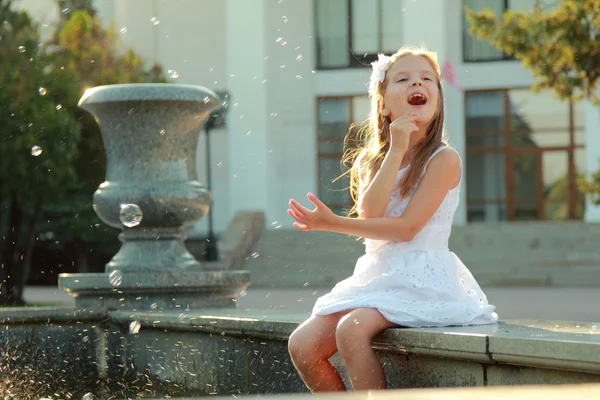 Linda chica joven sonriente en un hermoso vestido blanco pies de platija en la fuente y soplando burbujas de jabón — Foto de Stock