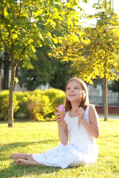 草の上に座って、アイスクリームを食べる白いドレスで甘い笑顔で魅力的な少女 — ストック写真