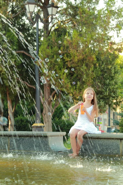 Mooi lachende meisje in een witte jurk bot voeten in de fontein en waait zeepbellen — Stockfoto