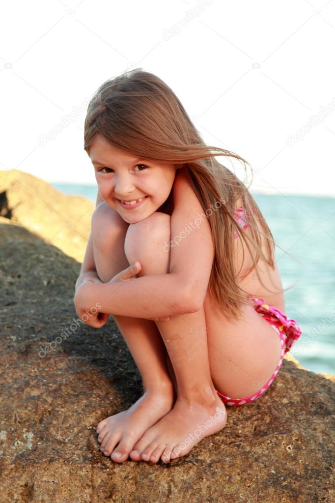 Cute little girl in a bathing suit sitting on a large rock by the sea.