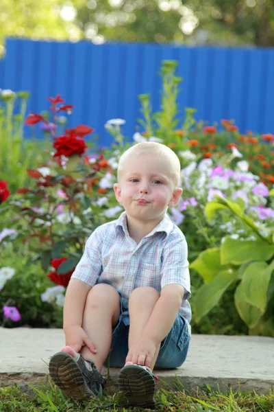 Glimlachend kleine blonde jongen speelt op de achtergrond van mooie bloemetjes buiten — Stockfoto