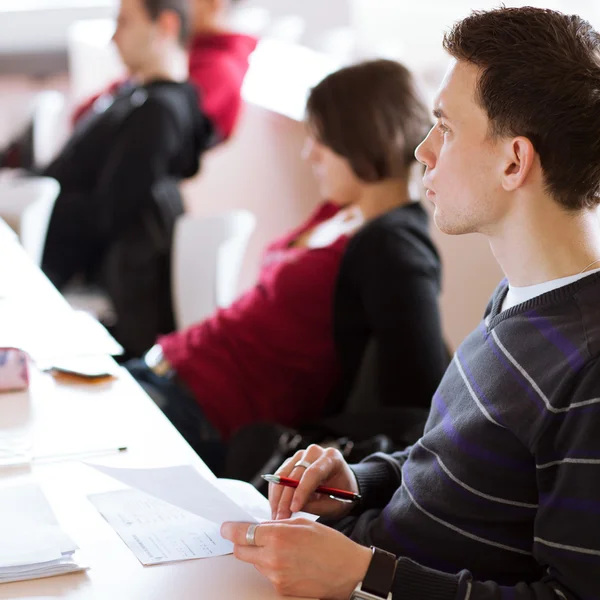 Ung, stilig hane högskolestudent som sitter i ett klassrum fullt — Stockfoto