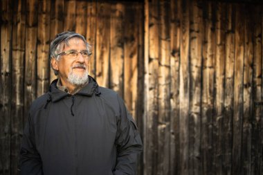 Kıdemli adam dışarıda gezmekten hoşlanıyor, yürüyüş yapıyor, güzel doğada yürüyor, ahşap bir dağ evinin önünde duruyor.