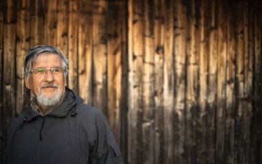 Kıdemli adam dışarıda gezmekten hoşlanıyor, yürüyüş yapıyor, güzel doğada yürüyor, ahşap bir dağ evinin önünde duruyor.