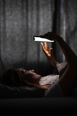 Yatakta elinde telefon tutan genç bir kadın, yorgun ve bitkin, mavi ışık gözlerini zorluyor, kafiyesini bozuyor.