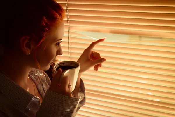 Femme avec café regarde à travers les stores tôt le matin — Photo