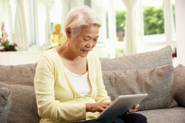 Senior Chinese Woman With Tablet Computer Whilst Relaxing On Sof clipart