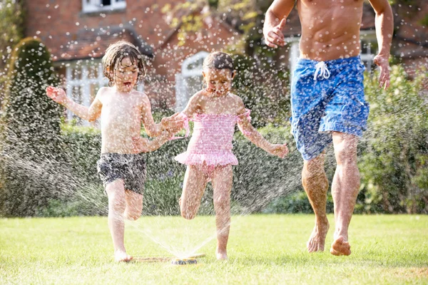 Vader en twee kinderen lopen via tuin sprinkler — Stockfoto