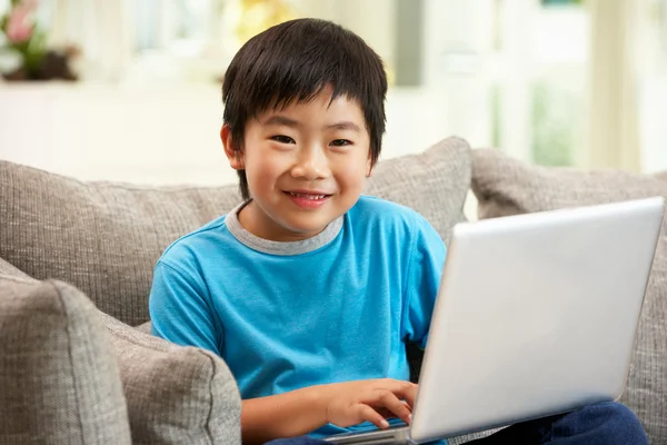 Menino chinês jovem usando laptop enquanto sentado no sofá em casa — Fotografia de Stock