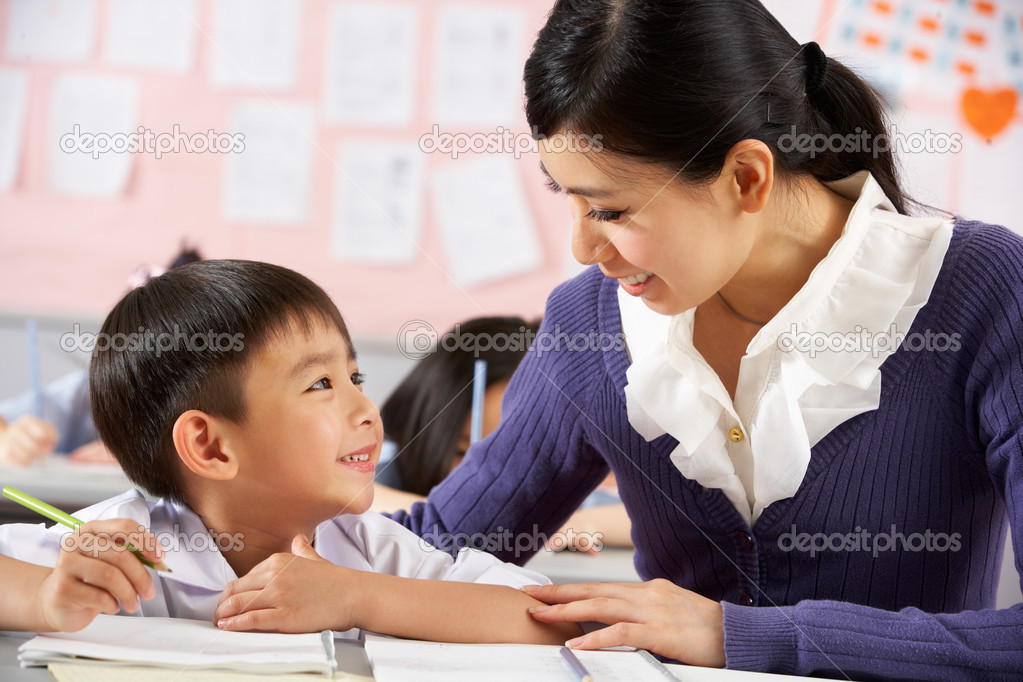 Teacher Helping Student Working At Desk In Chinese School Classr