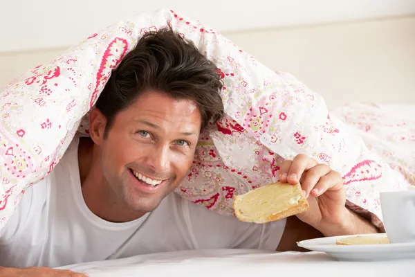Mann kuschelt sich unter Bettdecke und frühstückt — Stockfoto
