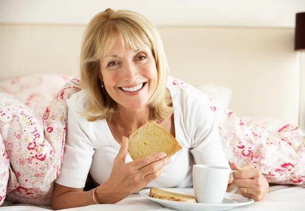Senior mujer acurrucado bajo edredón comiendo desayuno —  Fotos de Stock