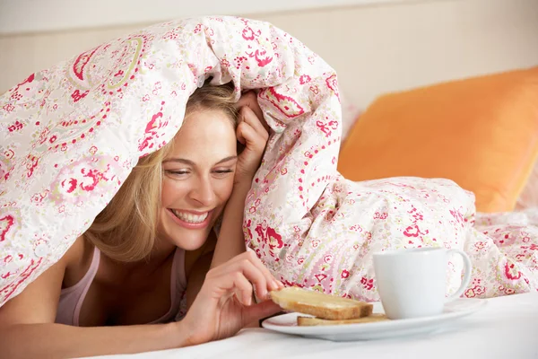 Hübsche Frau kuschelt sich unter Bettdecke und frühstückt — Stockfoto