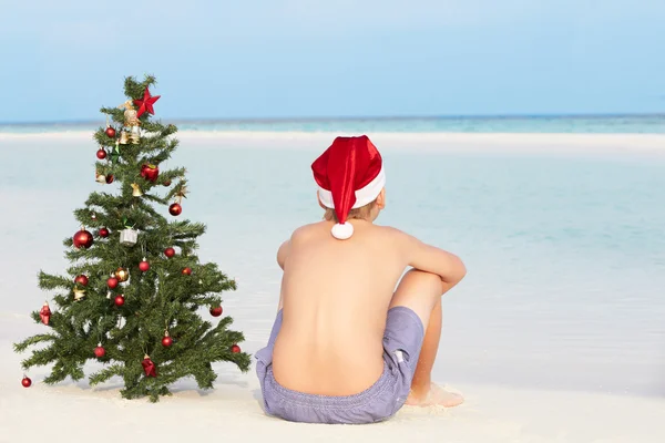Αγόρι καθιστός σε παραλία με το χριστουγεννιάτικο δέντρο και καπέλο — Φωτογραφία Αρχείου
