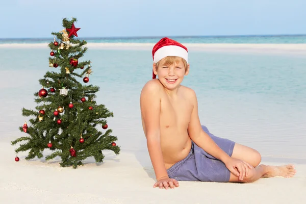 Pojken sitter på stranden med julgran och hatt — Stockfoto