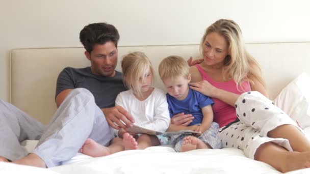 Kinderen zitten tussen de ouders in bed lezen van een boek samen. — Stockvideo