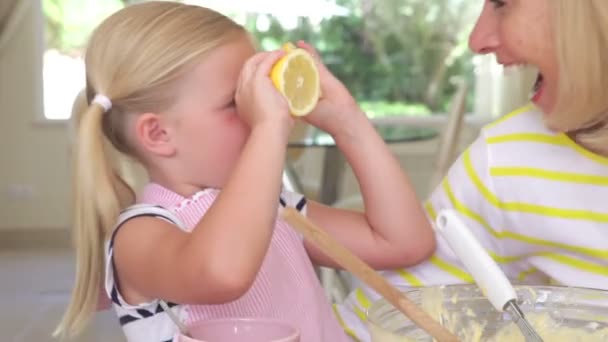 Grandmother and granddaughter sitting at kitchen table with ingredients as girl holds up lemon halves and makes funny face. — Stock Video