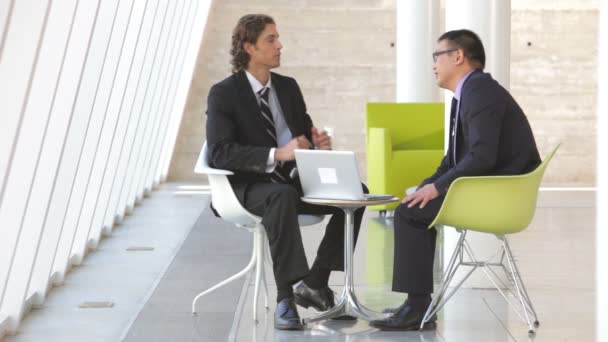 Два бизнесмена сидят за столом в современном офисе и смотрят на ноутбук вместе — стоковое видео