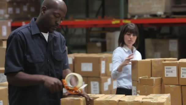Mannelijke fabriek werknemer verzegeling pakketten voor verzending als vrouwelijke collega controleert dozen tegen Klembord. — Stockvideo