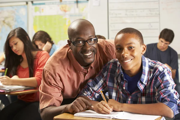 Teacher Helping Male Pupil Studying At Desk In Classroom Stock Picture