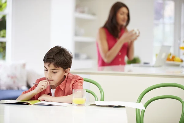 Sohn erledigt Hausaufgaben, während Mutter Laptop im Hintergrund benutzt — Stockfoto