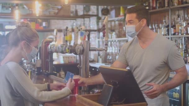 バーでマスクを身に着けている女性の顧客は 健康流行中の飲料のための非接触支払いを行う — ストック動画