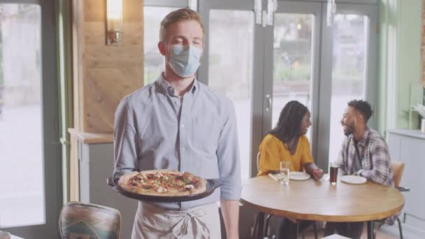 Porträtt Servitör Restaurang Bär Mask Som Serverar Pizza Till Par — Stockvideo