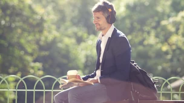 ビジネスマン読書やテイクアウトコーヒーと公園でヘッドフォンで聴く — ストック動画