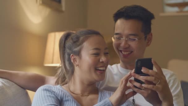 Sonriente Pareja Asiática Relajarse Sofá Casa Mirando Teléfono Móvil Noche — Vídeos de Stock