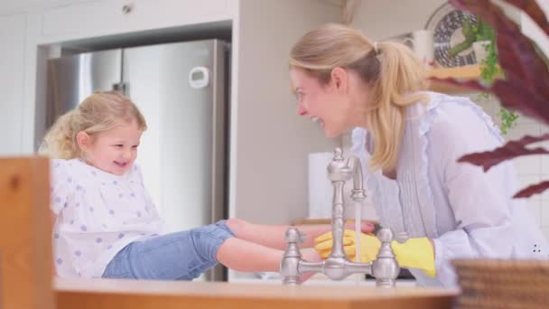 妈妈带着橡胶手套在厨房里待在家里 小女儿在厨房里玩得很开心 女儿把脚放在水池里 就像她们慢吞吞地洗澡一样 — 图库视频影像