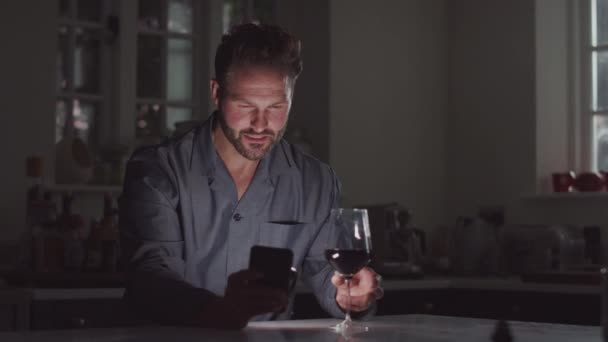 携帯電話を使って夜はワインを飲みながらキッチンに座るパイジャマ姿の熟女 — ストック動画