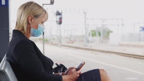 電車の駅のプラットフォーム上で顔マスクを身に着けている熟女は携帯電話でテキストメッセージを送信 スローモーションで撮影 — ストック動画