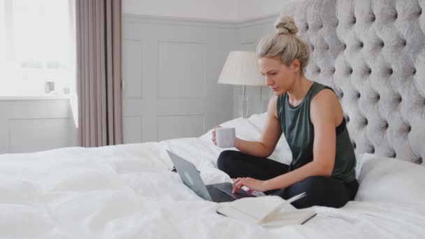 健康大流行病期间在家里工作的女商人坐在床上用笔记本电脑喝热饮的侧视图 慢镜头 — 图库视频影像