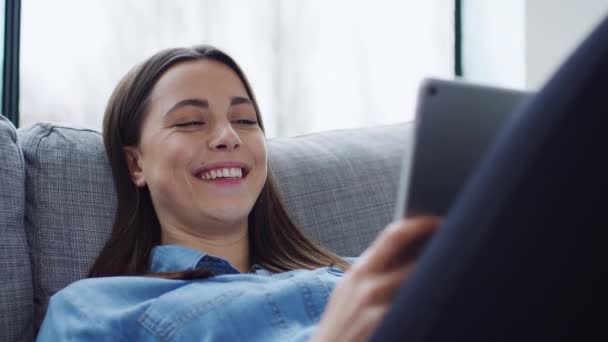 リラックス若いです女性に家に横たわっソファ見にデジタルタブレットと笑い 遅い動きで撮影 — ストック動画