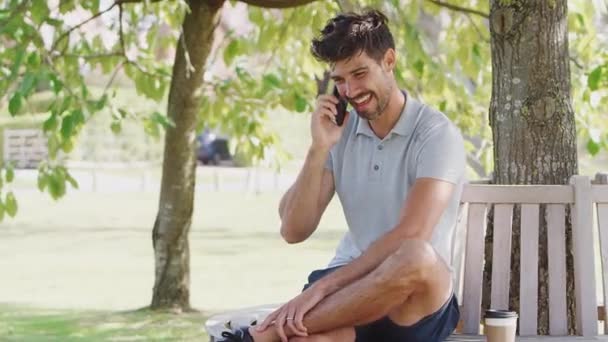 穿着短裤坐在树下公园的长椅上 悠闲地在手机上聊天的男人 慢镜头 — 图库视频影像