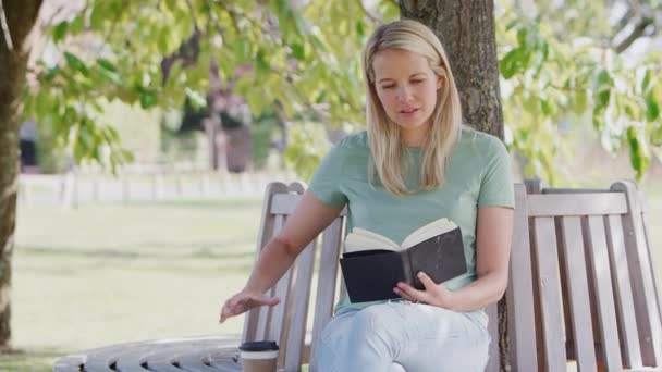 女人坐在公园的长椅上 在树下看书 喝着外卖咖啡 动作缓慢 — 图库视频影像