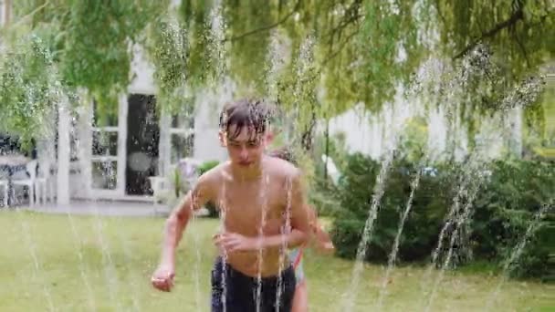 男の子と女の子でスイミング衣装夏に庭のスプリンクラーから水を通ってジャンプバックヤード スローモーションで撮影 — ストック動画