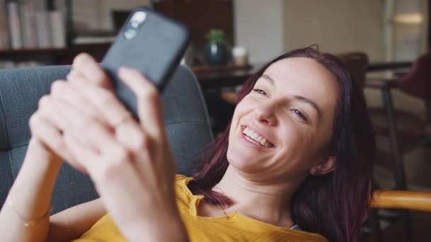 Χαμογελαστή Νεαρή Γυναίκα Ξαπλωμένη Στον Καναπέ Στο Σπίτι Κοιτάζοντας Κινητό — Αρχείο Βίντεο