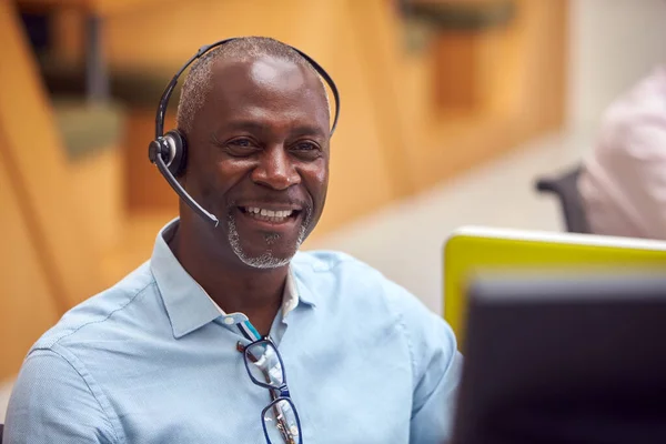 Portrait Of Mature Businessman Wearing Phone Headset Talking To Caller In Customer Services Centre