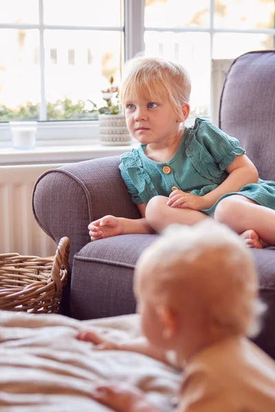 幼い女の子が家でソファに座ってテレビを見ている赤ちゃんの弟が前景で遊ぶ — ストック写真