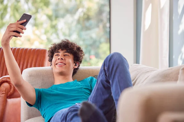 十几岁的男孩躺在沙发上在家用手机为社交媒体自拍 — 图库照片