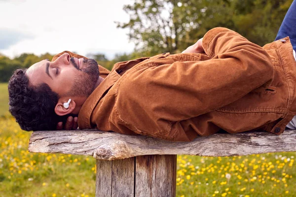 田舎のベンチに寝そべって音楽を聴いたり ワイヤレスイヤホンでポッドキャストを聴いたりする男 — ストック写真